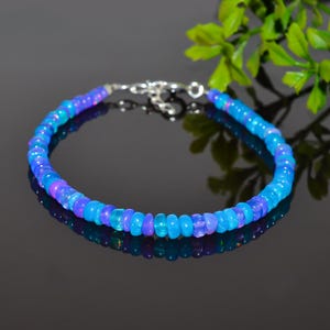 May include: A bracelet made of blue and purple opal beads, with a silver clasp. The beads are a mix of light and dark blues and purples. The bracelet is displayed on a reflective surface.