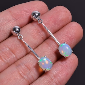 Op de afbeelding: Een paar zilveren oorhangers. Elke oorbel heeft een kleine, ronde knop, een lange, dunne staaf en een ovale opaal edelsteen. De opalen vertonen een kleurenspel met flitsen van groen, blauw en oranje. De oorbellen worden op een hand getoond.