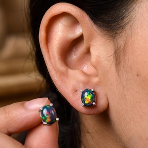 Könnte beinhalten: Ein Paar oval geformte schwarze Opal-Ohrstecker, gefasst in silberfarbene Metallkrappen. Ein Ohrstecker sitzt im Ohrläppchen, während der andere zwischen den Fingern gehalten wird und die irisierenden roten, orangen und grünen Blitze im Stein zeigt.