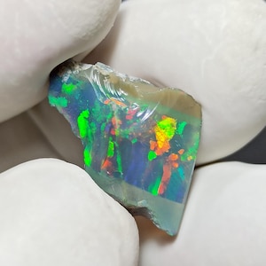 Peut inclure: Une opale vibrante aux couleurs variées, notamment le vert, le bleu et l'orange. La pierre brute présente un jeu de lumière, mettant en valeur sa beauté naturelle. L'opale est tenue par une main gantée, soulignant ses caractéristiques uniques.
