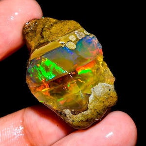 May include: A rough opal gemstone with vibrant colors of green, orange, and blue. The stone has a yellow-brown matrix and a fractured surface, showcasing its natural, raw form. The opal is held in a hand against a black background.