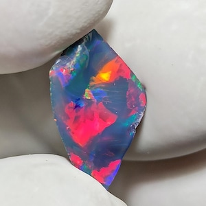 May include: A vibrant, raw opal gemstone with a dark base color. The stone displays a play of color with flashes of red, orange, green, and blue. The opal is held by a gloved hand, showcasing its natural, uncut form.