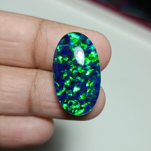 Puede incluir: Una piedra preciosa de ópalo negro de forma ovalada con destellos verdes y azules vibrantes. La piedra pulida muestra un juego de color, con una base oscura y tonos brillantes y cambiantes. El ópalo se sostiene en una mano.