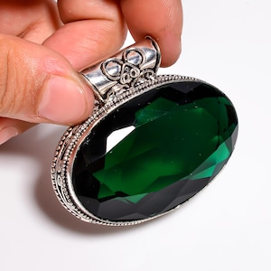 Pode incluir: Pingente oval com uma grande pedra preciosa verde esmeralda facetada. A pedra é engastada em uma moldura de metal prateado com detalhes intrincados. O pingente é segurado por uma mão, mostrando o design da joia.