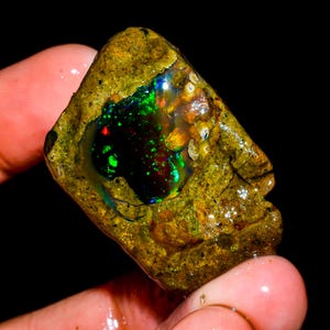 May include: A rough-cut opal gemstone with a dark, iridescent center displaying flashes of green, red, and blue. The opal is encased in a rough, golden-brown matrix. The stone is held up against a black background.