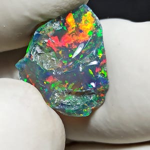 May include: A vibrant, raw opal gemstone with a dark base color. The opal displays a play of color, including red, orange, green, and blue hues. The stone is irregularly shaped and appears to be freshly mined.