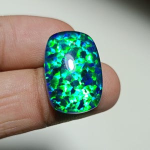 Könnte beinhalten: Ein polierter Opal-Edelstein mit rechteckiger Form und abgerundeten Ecken. Der Stein zeigt ein Mosaik aus leuchtenden Grün- und tiefblauen Farben mit einer reflektierenden Oberfläche. Der Opal wird zwischen Fingern gehalten.