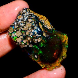 May include: A rough, uncut opal gemstone with a dark, iridescent body. The opal displays flashes of green, blue, and red, with a matrix of tan and brown inclusions. The stone is held in a hand, showcasing its natural, raw beauty.