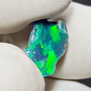 May include: A vibrant, raw opal gemstone with a striking play of color. The stone displays vivid flashes of green and blue, with hints of yellow. The rough-cut edges and natural texture of the opal are visible.