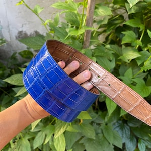 May include: A blue alligator leather belt with a brown leather lining. The belt is folded in half and held in a hand.