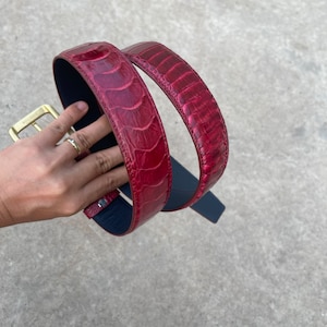 Op de afbeelding: Een rode leren riem met een textuurpatroon. De riem heeft een goudkleurige gesp en een donkerblauwe onderkant. De riem wordt in een gebogen positie getoond.