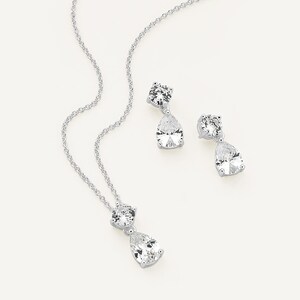 Könnte beinhalten: Ein silberfarbenes Schmuckset mit einer Halskette und Ohrringen. Die Halskette hat eine Kette mit einem runden und einem tropfenförmigen Anhänger aus klarem Stein. Die Ohrringe haben ein ähnliches Design mit runden und tropfenförmigen Steinen.