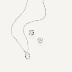 May include: A silver necklace with a round, clear gemstone pendant and matching stud earrings. The jewelry set is displayed on a white background, showcasing the sparkle of the stones and the delicate chain.