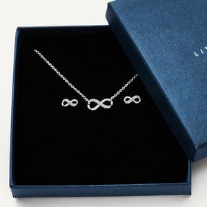 Puede incluir: Un conjunto de collar y pendientes plateados, con símbolos de infinito, presentado en una caja de regalo azul oscuro con interior negro. El collar y los pendientes están adornados con pequeños adornos brillantes.