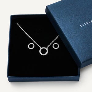Könnte beinhalten: Ein silbernes Halsketten- und Ohrring-Set mit einem runden Anhänger und passenden Ohrringen, präsentiert in einer dunkelblauen Geschenkbox. Der Schmuck wird auf einem schwarzen Samtinnenraum präsentiert, mit dem Wort "LITTLE" auf der Box.