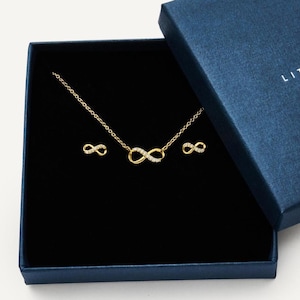 May include: A gold-coloured infinity symbol necklace and matching earrings set, presented in a dark blue gift box. The jewellery features small, sparkling accents. The box is open, revealing the jewellery against a black interior.