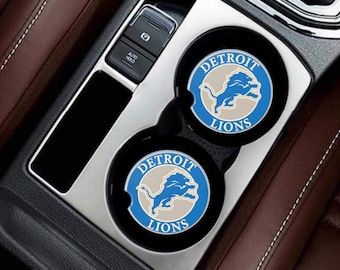 Puede incluir: Dos posavasos negros para coche con el logotipo de los Detroit Lions, con un león azul y plateado sobre un fondo azul claro y blanco, colocados en los portavasos de un coche. La consola es negra y plateada.