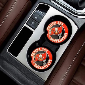 Puede incluir: Dos posavasos negros para coche con el logotipo de los Tampa Bay Buccaneers. El logotipo presenta una bandera roja con una calavera y espadas cruzadas, rodeada por las palabras "TAMPA BAY BUCCANEERS" en blanco. Los posavasos están en los portavasos de un coche.