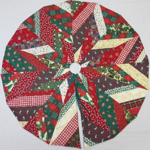 May include: Circular Christmas tree skirt with a patchwork design. The skirt features red, green, and white fabrics with holiday patterns like poinsettias, candy canes, and plaid. The center has a white circle and a split opening.