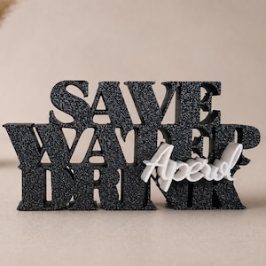 Könnte beinhalten: Schwarz-weißes Dekoschild mit der Aufschrift "SAVE WATER DRINK Aperol". Das Schild besteht aus strukturiertem Material und wird vor einem neutralen Hintergrund präsentiert. Eine Vase mit getrockneten Pflanzen befindet sich im Hintergrund.
