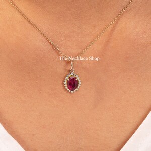 May include: A gold chain necklace with a pendant featuring a large oval ruby surrounded by a halo of smaller diamonds. The necklace is worn by a person with a white shirt. The text "The Necklace Shop" is visible in the image.