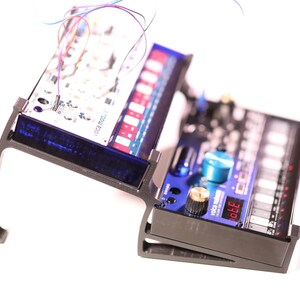 以下が含まれることがあります： 電子音楽機器を置くための2段式の黒いスタンド。 上段には、前面に「volca modular」と書かれた青と白のシンセサイザーが置かれています。 下段には、前面に「volca sample」と書かれた黒と青のシンセサイザーが置かれています。