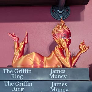May include: A 3D-printed figurine in shades of orange and red, posed atop a stack of books. The figurine is of a person with a flame-like head, holding a small flame. The books are titled "The Griffin Ring" and "The Last Paladin".