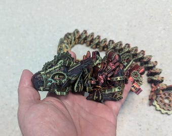 May include: A 3D printed dragon made of metal gears and cogs. The dragon is green, brown, and red with a metallic sheen. The dragon is curled up in a sleeping position.