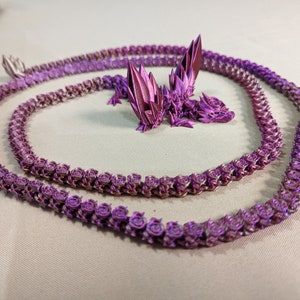 May include: Two purple origami dragon necklaces made of paper. The necklaces are in the shape of a dragon and are made of many small, folded pieces of paper.