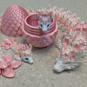 Puede incluir: Una figurita de drag&oacute;n blanco con detalles de flores de cerezo rosas. El drag&oacute;n est&aacute; enroscado en un c&iacute;rculo, con la cabeza apoyada en la cola. El drag&oacute;n est&aacute; hecho de un pl&aacute;stico blanco liso y tiene un acabado brillante. Las flores de cerezo est&aacute;n hechas de un pl&aacute;stico rosa suave y tienen un acabado mate.