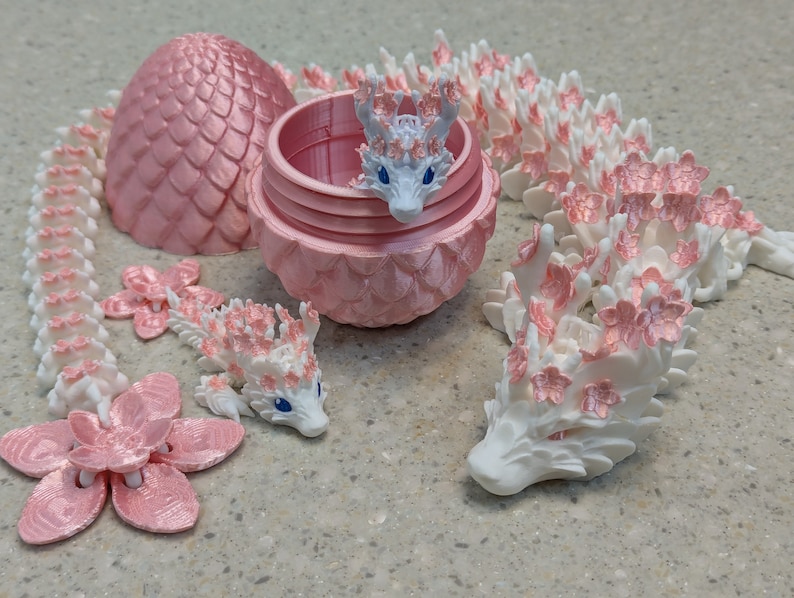 Puede incluir: Una figurita de drag&oacute;n blanco con detalles de flores de cerezo rosas. El drag&oacute;n est&aacute; enroscado en un c&iacute;rculo, con la cabeza apoyada en la cola. El drag&oacute;n est&aacute; hecho de un pl&aacute;stico blanco liso y tiene un acabado brillante. Las flores de cerezo est&aacute;n hechas de un pl&aacute;stico rosa suave y tienen un acabado mate.