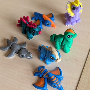 May include: A collection of seven small, articulated dinosaur figurines in various colors including purple with a yellow frill, green with an orange crest, blue with a tan stripe, dark green with red spikes, grey, and two blue and orange flying reptiles.