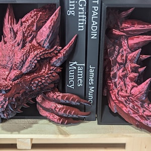May include: Red dragon-shaped bookends. One bookend features the dragon's head, the other its tail. The bookends are placed on a wooden shelf, with black books between them. The books have white text that reads "PALADIN" and "Griffin" and the author's name.