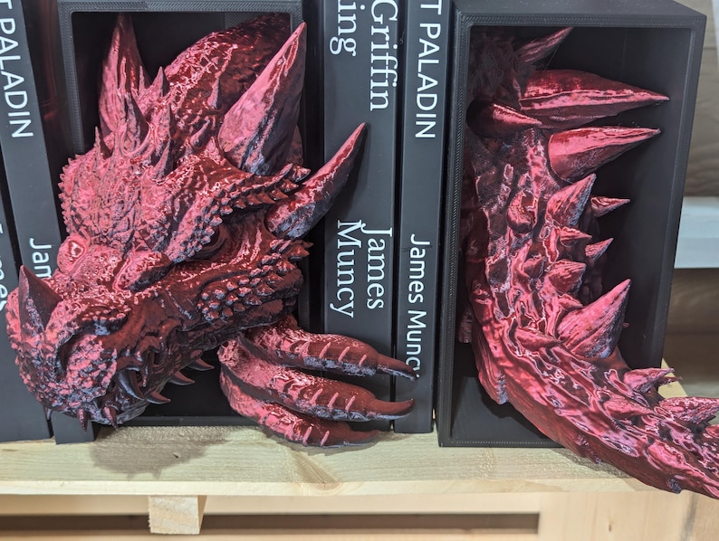 May include: Red dragon-shaped bookends. One bookend features the dragon's head, the other its tail. The bookends are placed on a wooden shelf, with black books between them. The books have white text that reads "PALADIN" and "Griffin" and the author's name.
