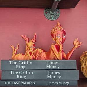 May include: A 3D-printed sculpture of two figures with flame-like hair and features, lying atop three books. The books are titled "The Griffin Ring" and "The Last Paladin" by James Muncy. The sculpture is in shades of red and gold.