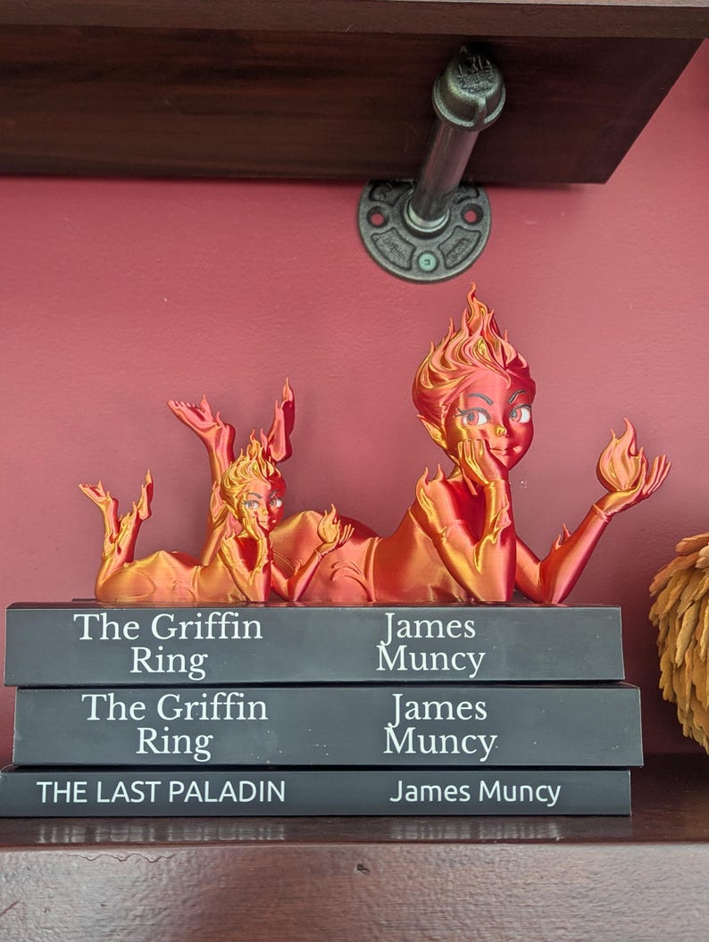 May include: A 3D-printed sculpture of two figures with flame-like hair and features, lying atop three books. The books are titled "The Griffin Ring" and "The Last Paladin" by James Muncy. The sculpture is in shades of red and gold.