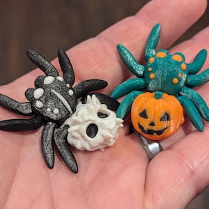May include: Two Halloween-themed spider figurines. One spider is black with white spots and a white skull head. The other spider is green with orange spots and is holding a small orange pumpkin with a jack-o'-lantern face.