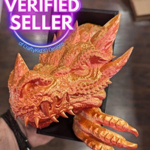 May include: A 3D-printed orange dragon head with sharp teeth and textured scales. The dragon head is held in a black box. The image includes a circular "VERIFIED SELLER" graphic with the text "of CraftyKid3D Designs".