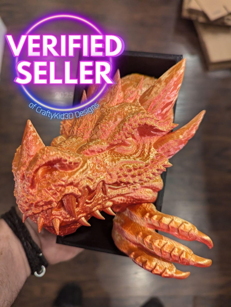 May include: A 3D-printed orange dragon head with sharp teeth and textured scales. The dragon head is held in a black box. The image includes a circular "VERIFIED SELLER" graphic with the text "of CraftyKid3D Designs".