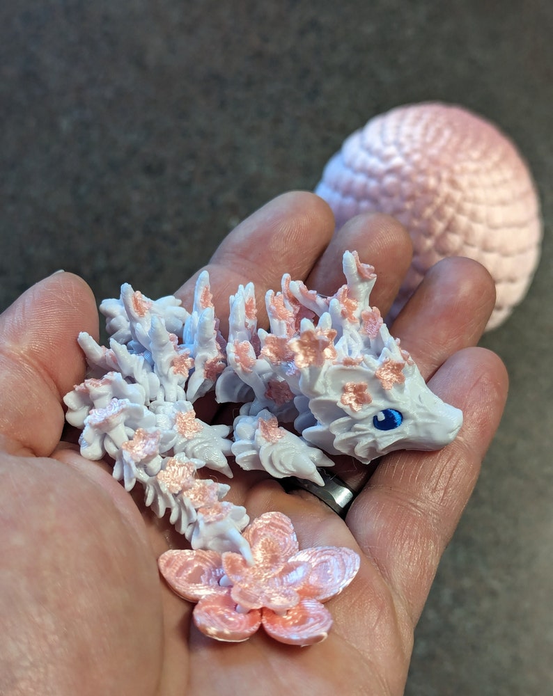 Puede incluir: Una peque&ntilde;a figura de drag&oacute;n blanca y rosa con una cola de flor. El drag&oacute;n tiene un ojo azul y est&aacute; cubierto de peque&ntilde;os detalles de flores rosas.