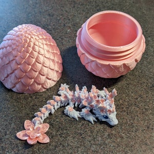 Puede incluir: Un huevo de drag&oacute;n rosa con una tapa extra&iacute;ble, que revela un peque&ntilde;o drag&oacute;n blanco con detalles rosas en el interior. El drag&oacute;n tiene un ojo azul y una flor rosa en la cola.