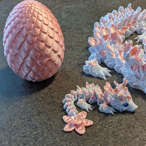 Puede incluir: Tres figuras de drag&oacute;n impresas en 3D de color rosa y blanco. El drag&oacute;n m&aacute;s grande est&aacute; enroscado con la cabeza mirando hacia la derecha. Los dos dragones m&aacute;s peque&ntilde;os est&aacute;n mirando hacia la izquierda. El drag&oacute;n m&aacute;s grande tiene un dise&ntilde;o floral rosa y blanco. Los dos dragones m&aacute;s peque&ntilde;os tienen un dise&ntilde;o floral rosa y blanco. Los dragones est&aacute;n sentados sobre una superficie gris.