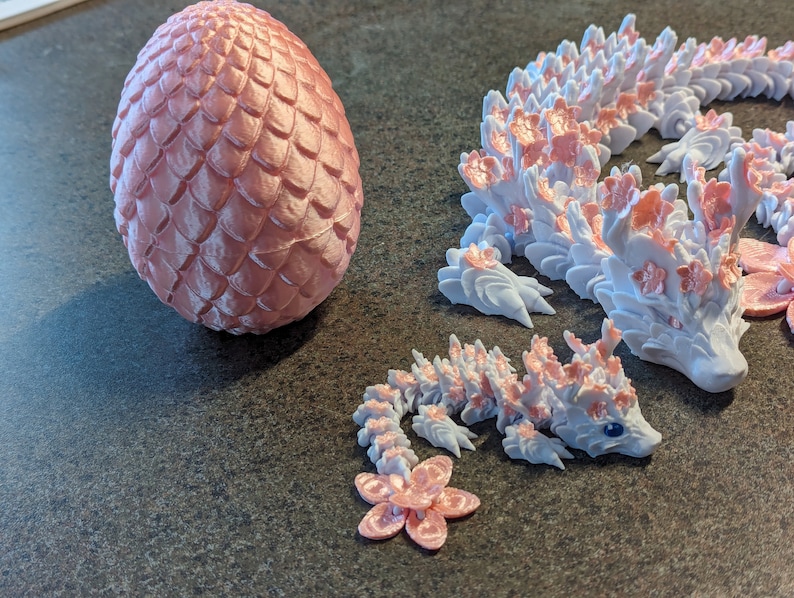 Puede incluir: Tres figuras de drag&oacute;n impresas en 3D de color rosa y blanco. El drag&oacute;n m&aacute;s grande est&aacute; enroscado con la cabeza mirando hacia la derecha. Los dos dragones m&aacute;s peque&ntilde;os est&aacute;n mirando hacia la izquierda. El drag&oacute;n m&aacute;s grande tiene un dise&ntilde;o floral rosa y blanco. Los dos dragones m&aacute;s peque&ntilde;os tienen un dise&ntilde;o floral rosa y blanco. Los dragones est&aacute;n sentados sobre una superficie gris.
