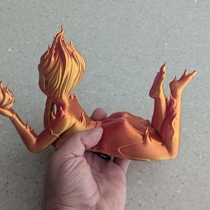 May include: A 3D-printed sculpture of a reclining figure with flames for hair and body details. The figure is holding a flame in one hand. The sculpture is in shades of orange and gold.