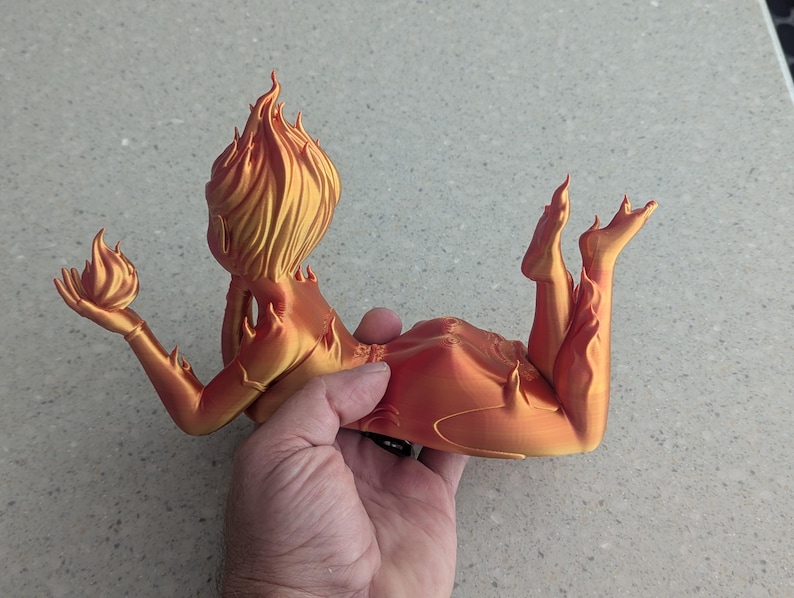 May include: A 3D-printed sculpture of a reclining figure with flames for hair and body details. The figure is holding a flame in one hand. The sculpture is in shades of orange and gold.