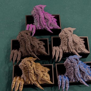 May include: Five colorful, textured dragon busts in black display boxes. The dragons are in shades of purple, black, gray, gold, and blue. Each dragon has detailed scales and claws, with a textured, rough appearance. The busts are arranged on a green surface.