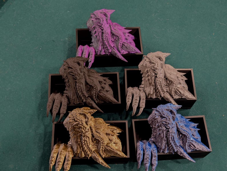 May include: Five colorful, textured dragon busts in black display boxes. The dragons are in shades of purple, black, gray, gold, and blue. Each dragon has detailed scales and claws, with a textured, rough appearance. The busts are arranged on a green surface.