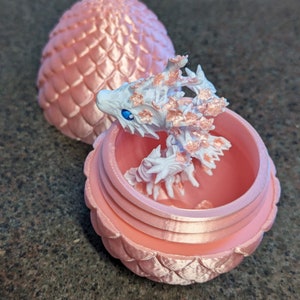 Puede incluir: Un huevo de drag&oacute;n rosa con un drag&oacute;n blanco saliendo de &eacute;l. El huevo est&aacute; hecho de pl&aacute;stico y tiene una superficie texturizada. El drag&oacute;n tambi&eacute;n est&aacute; hecho de pl&aacute;stico y tiene un esquema de color blanco y rosa.