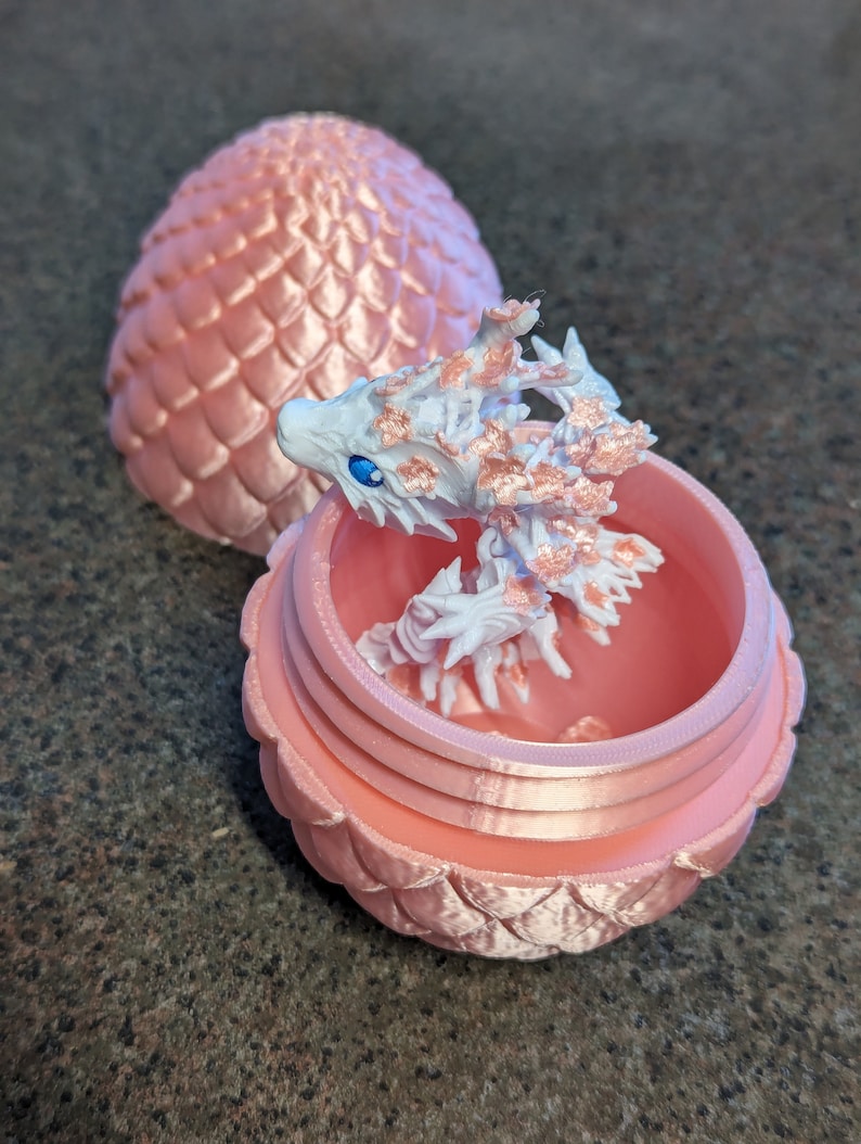 Puede incluir: Un huevo de drag&oacute;n rosa con un drag&oacute;n blanco saliendo de &eacute;l. El huevo est&aacute; hecho de pl&aacute;stico y tiene una superficie texturizada. El drag&oacute;n tambi&eacute;n est&aacute; hecho de pl&aacute;stico y tiene un esquema de color blanco y rosa.