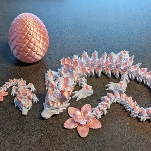 Puede incluir: Un conjunto de tres figuras de drag&oacute;n impresas en 3D de color rosa y blanco. El drag&oacute;n m&aacute;s grande est&aacute; enroscado en un c&iacute;rculo, el drag&oacute;n mediano est&aacute; tumbado y el drag&oacute;n m&aacute;s peque&ntilde;o est&aacute; de pie sobre sus patas traseras. Los dragones est&aacute;n todos decorados con flores rosas.